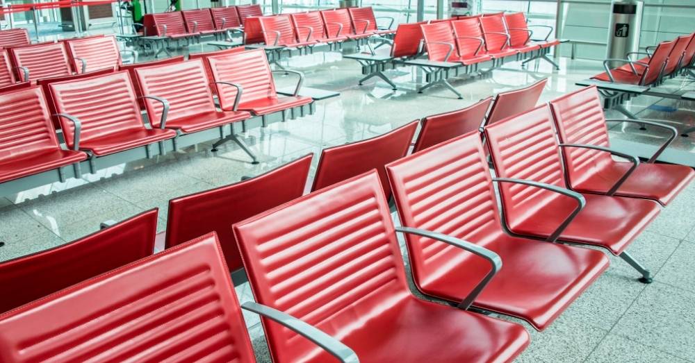 Several rows of empty, shiny red waiting chairs are shown in an airport or large waiting room.