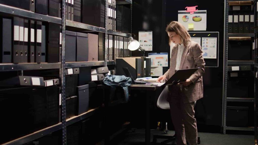 Woman reviewing files in a physical records storage room with office shelves and binders