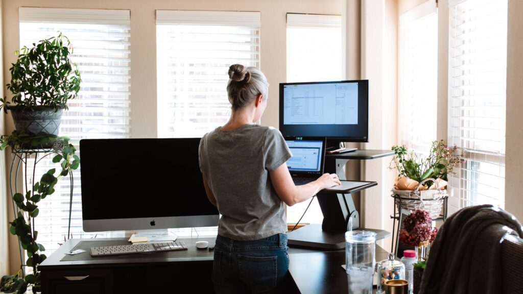 Woman standing while using a laptop and monitor in a bright, modern office space, exemplifying standing desk ergonomics