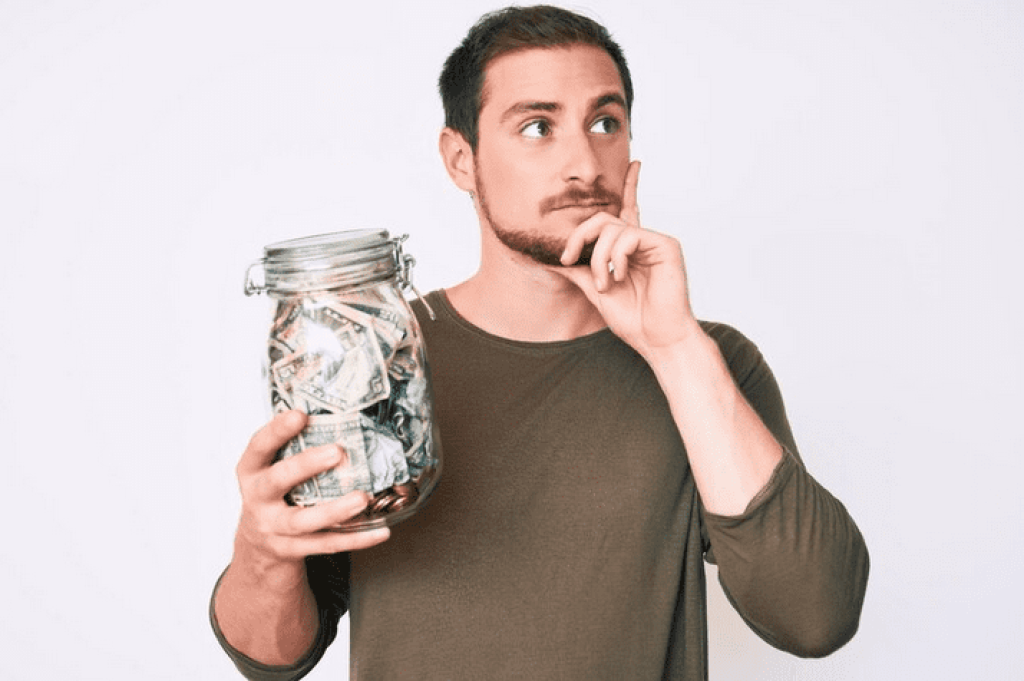 Young handsome man holding jar with savings serious face thinking about question with hand on chin thoughtful about confusing idea